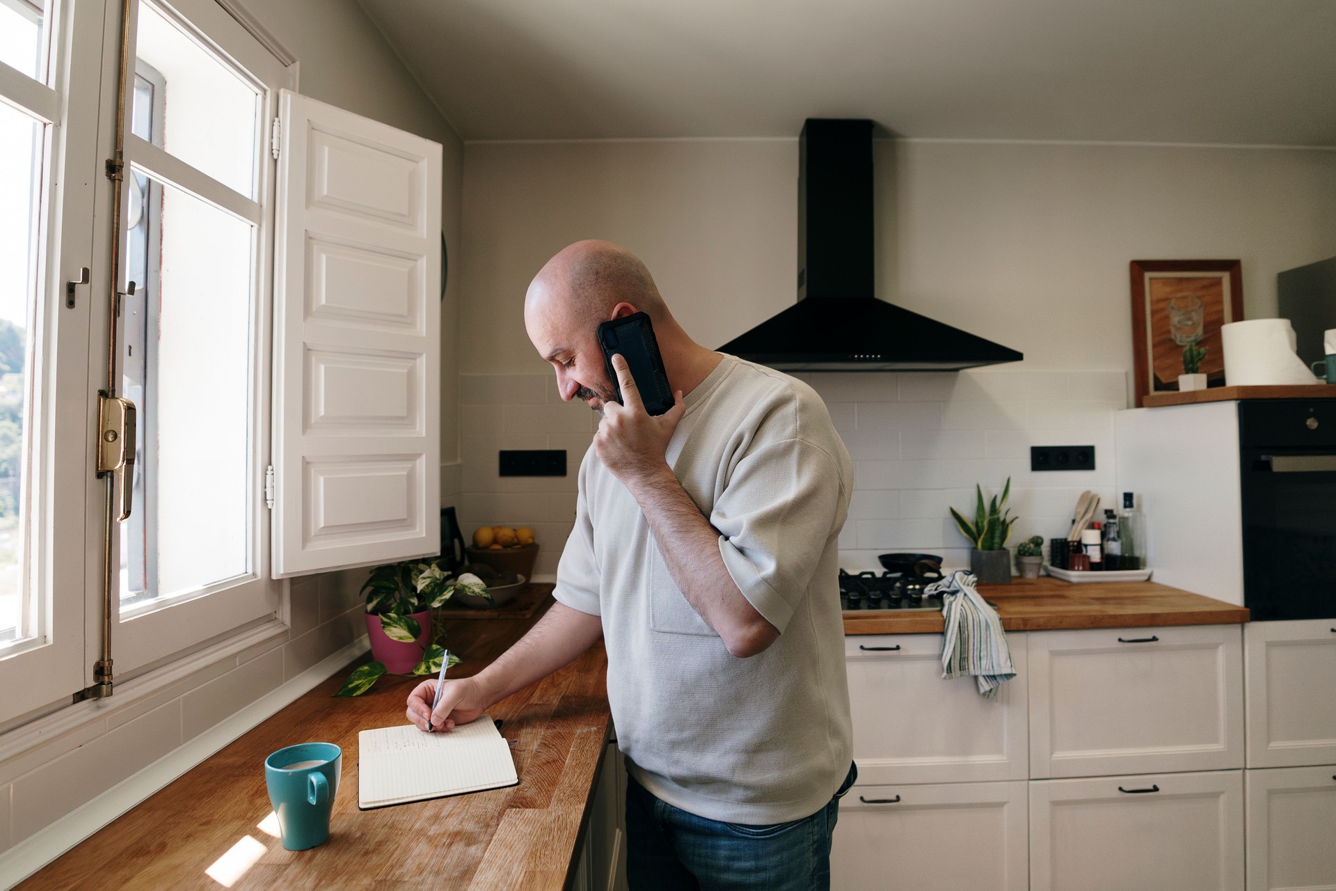 Hombre en la cocina escribiendo en el cuaderno mientras habla por teléfono celular
